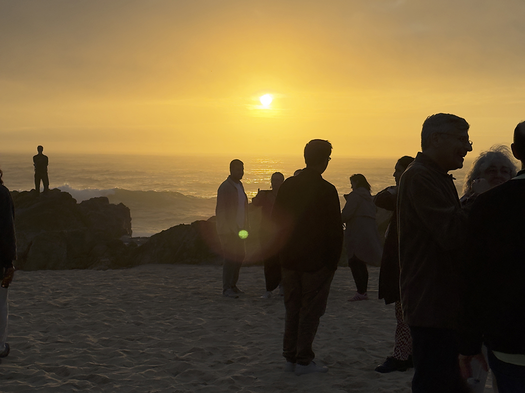 People stand and talk on a beach at sunset, their silhouettes outlined against the glowing sky and shimmering ocean.