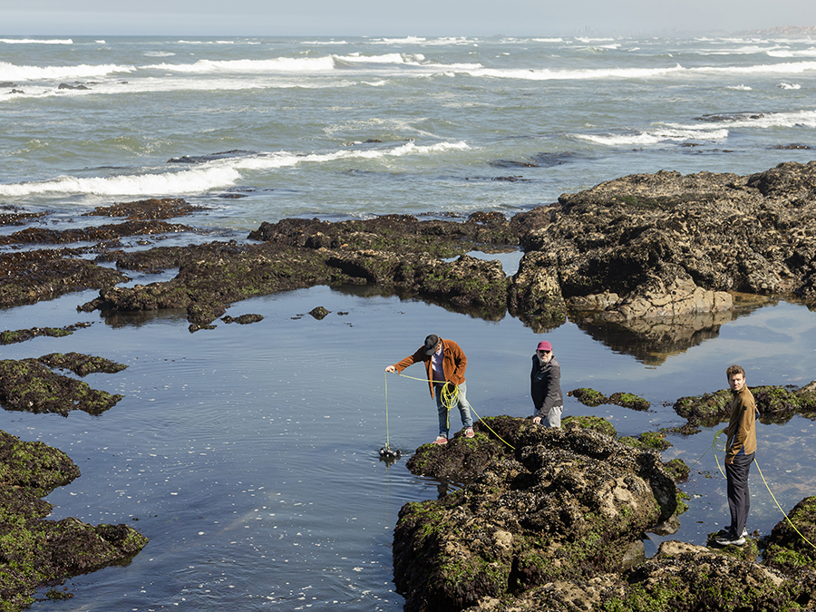 Three people stand on rocky tide pools by the ocean, using ropes to lower ROV equipment into the water.